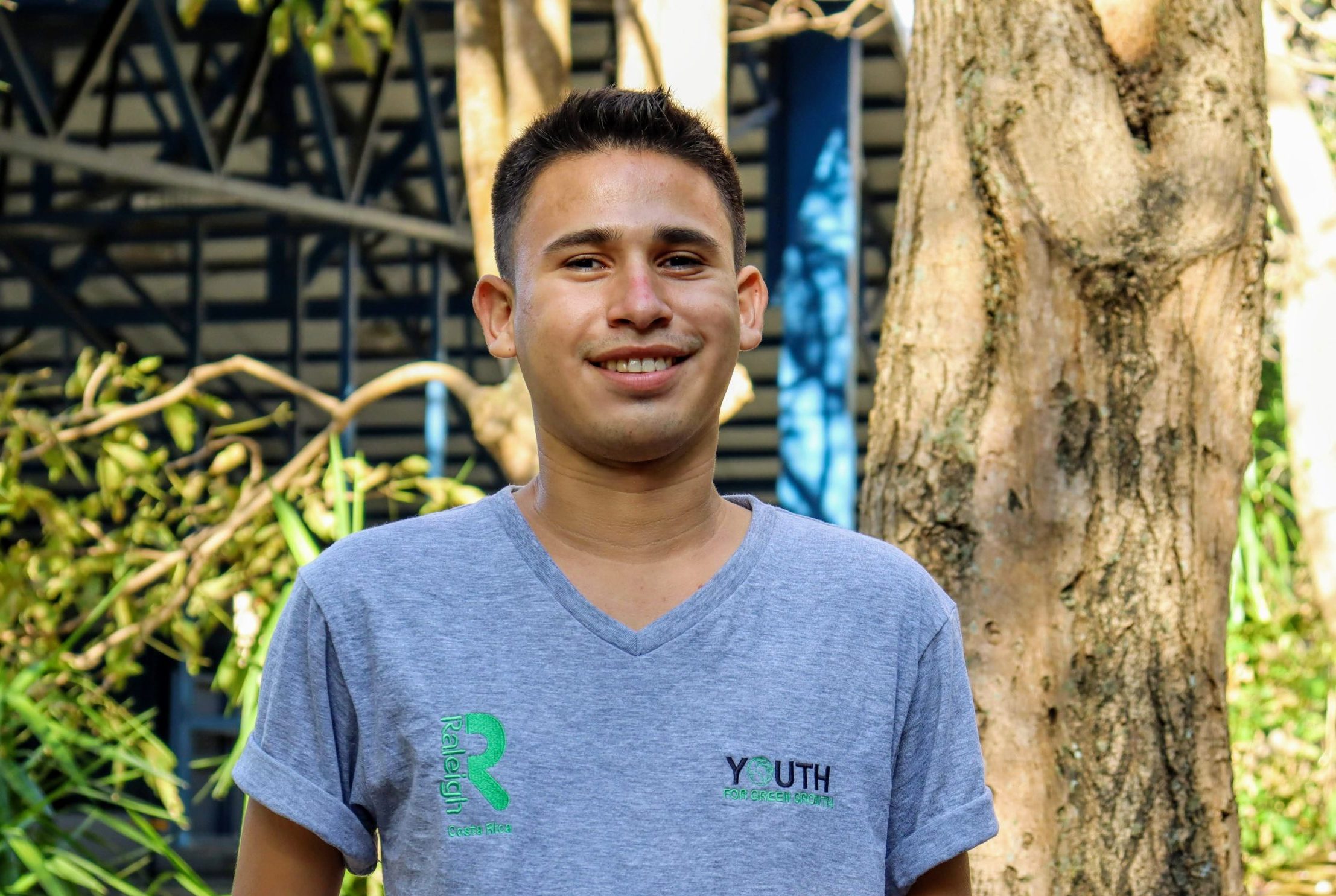 Young man smiling outdoors wearing a grey V-neck T-shirt with logos from Raleigh Costa Rica and Youth for Green Growth.